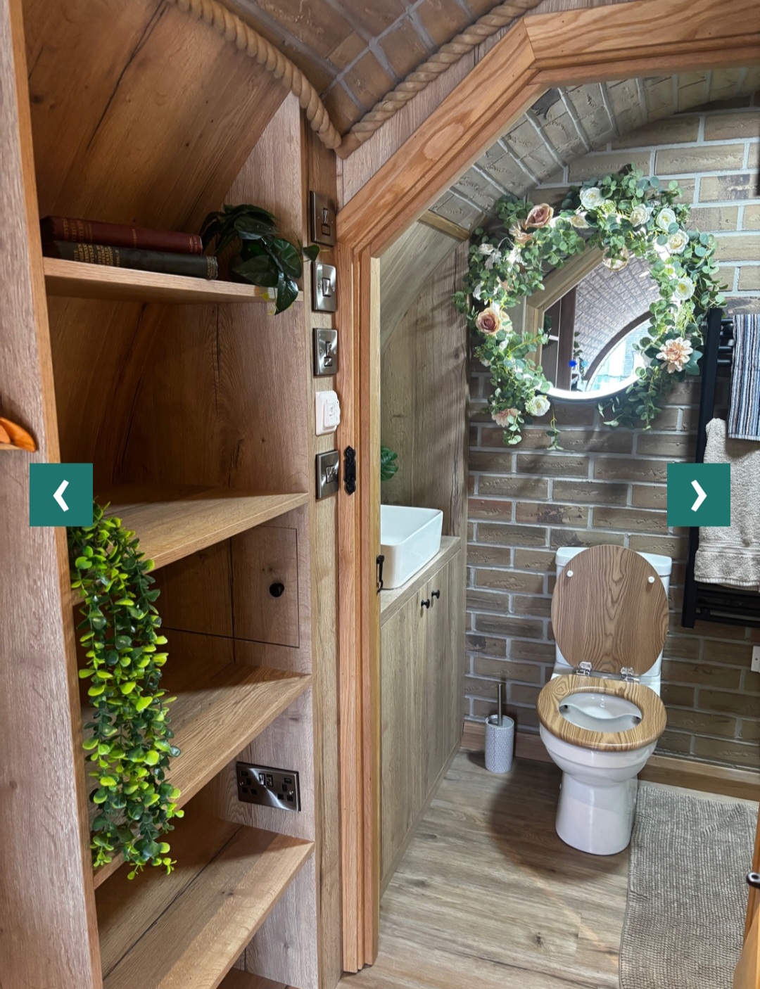 Bathroom detail — oak shelving with books and eucalyptus, arched doorway to the toilet with a floral wreath mirror.