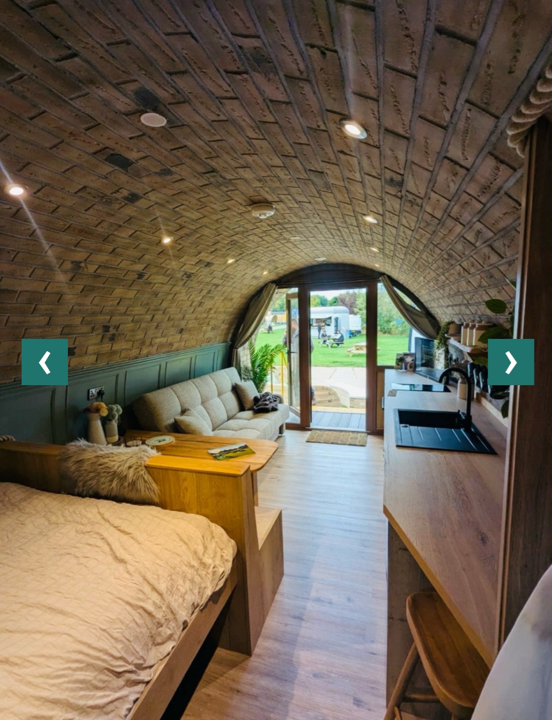 Interior looking from the bed through the living area to the open door — sofa on the left, kitchen on the right, pasture beyond.