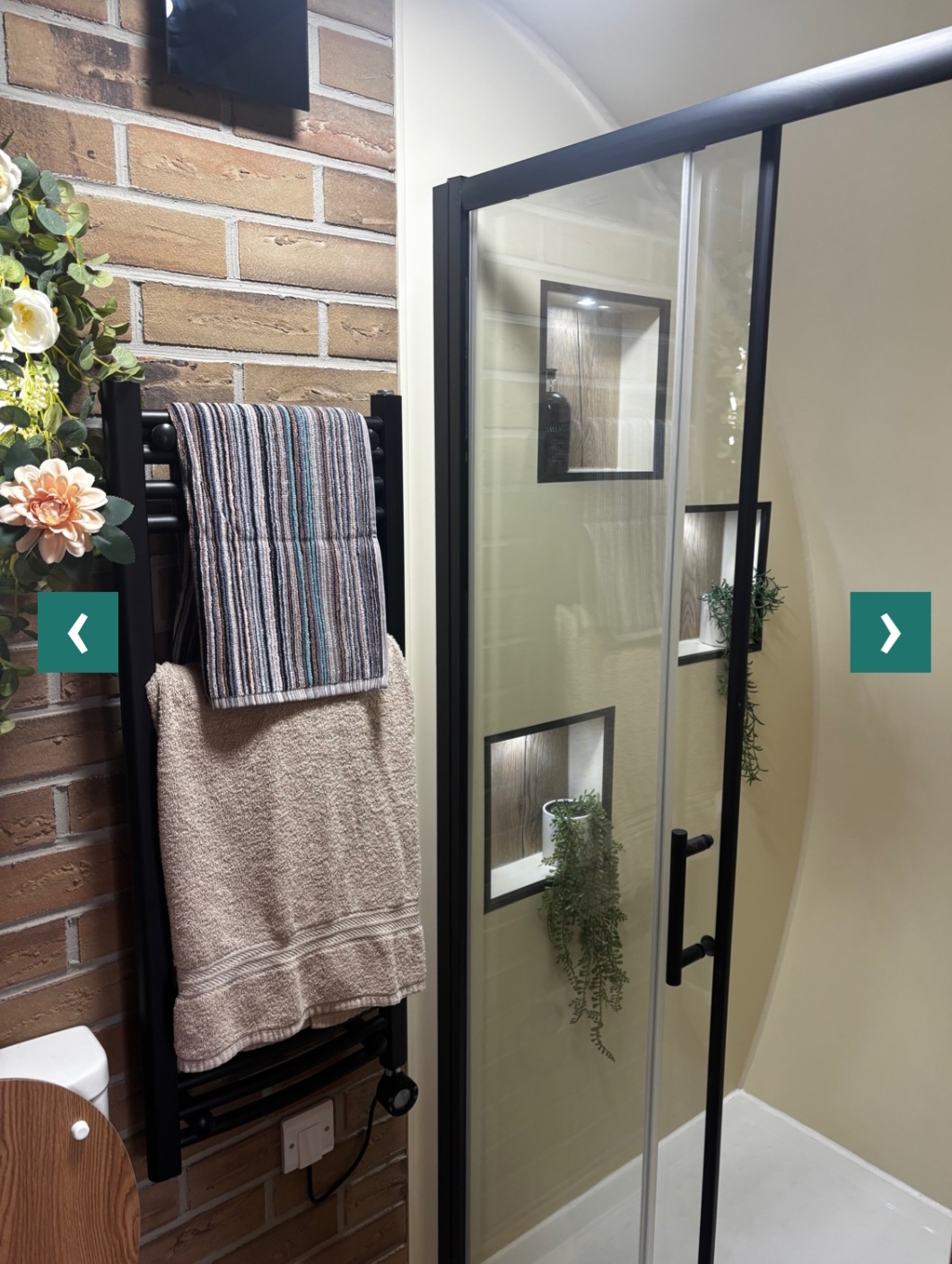 Shower detail — glass shower door, matte-black towel radiator, warm brick wall.